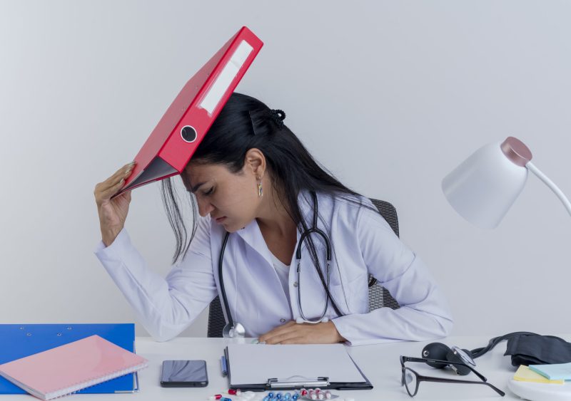 stressed young female doctor wearing medical robe and stethoscope sitting at desk with medical tools putting hand on desk touching head with folder with closed eyes isolated on white background